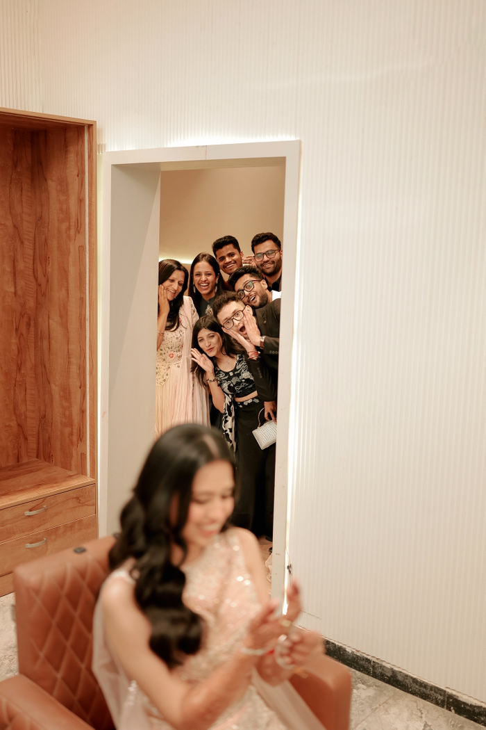 Cinematic candid photograph of a bride in soft focus as her friends peek through a doorway during wedding preparations. | Studio Memory Lane