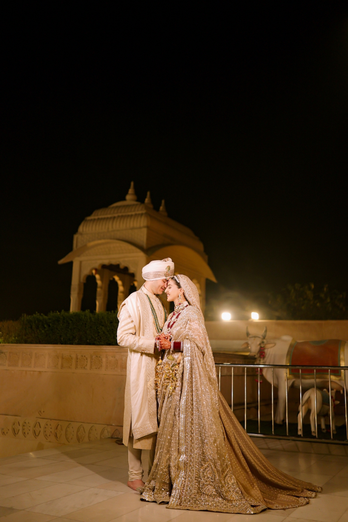 Cinematic wedding portrait of a bride and groom in elegant ivory and gold attire, standing close together beneath the warm evening lights of a wedding venue.