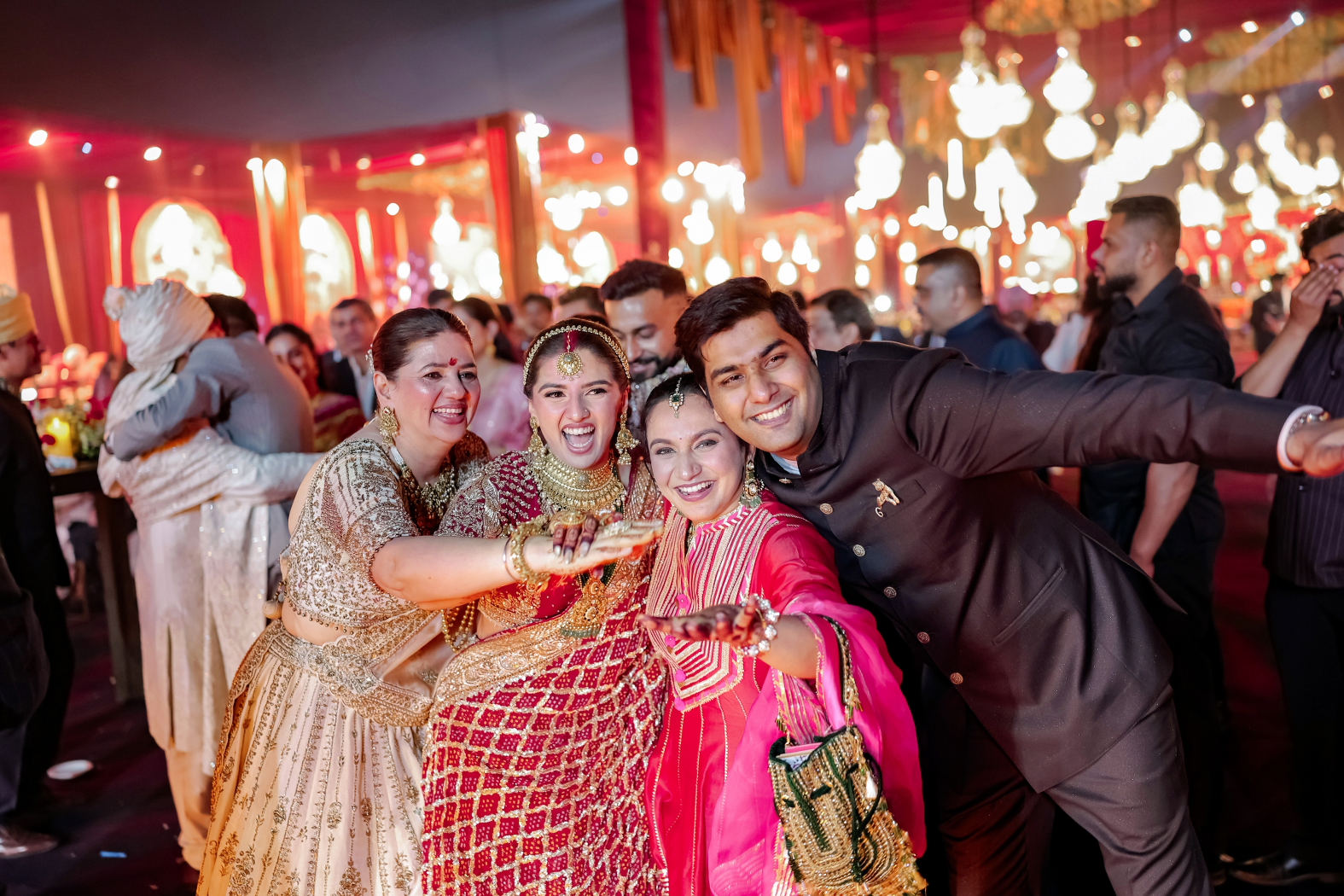 A joyful wedding reception portrait of the bride in a red bridal lehenga posing with friends and family, surrounded by guests and warm, festive lighting.