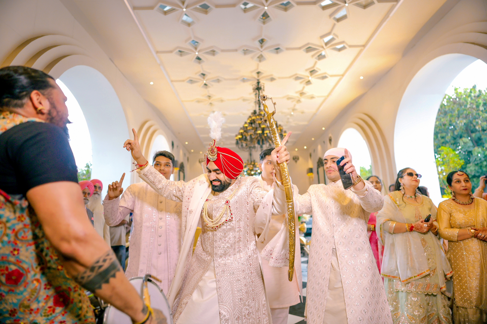 Groom in a red turban and ivory sherwani dancing with his family during a lively wedding baraat inside a beautifully decorated hall.