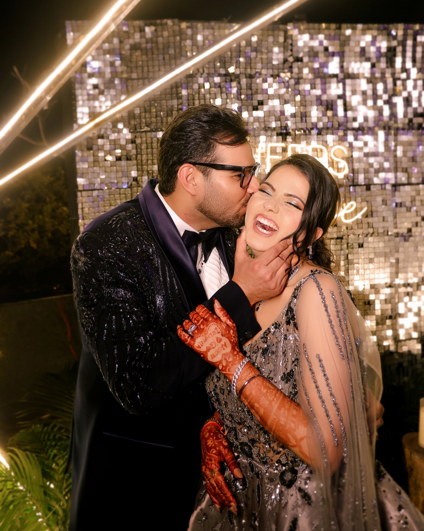 A romantic wedding reception portrait of a groom kissing his bride on the cheek as she laughs, showcasing her intricate mehndi, shimmering silver lehenga, and joyful candid expression against a glittering backdrop.