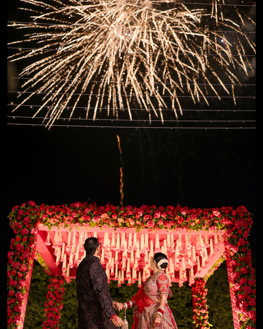 A newlywed couple standing beneath a floral wedding canopy as golden fireworks illuminate the night sky, creating a cinematic and romantic wedding portrait.