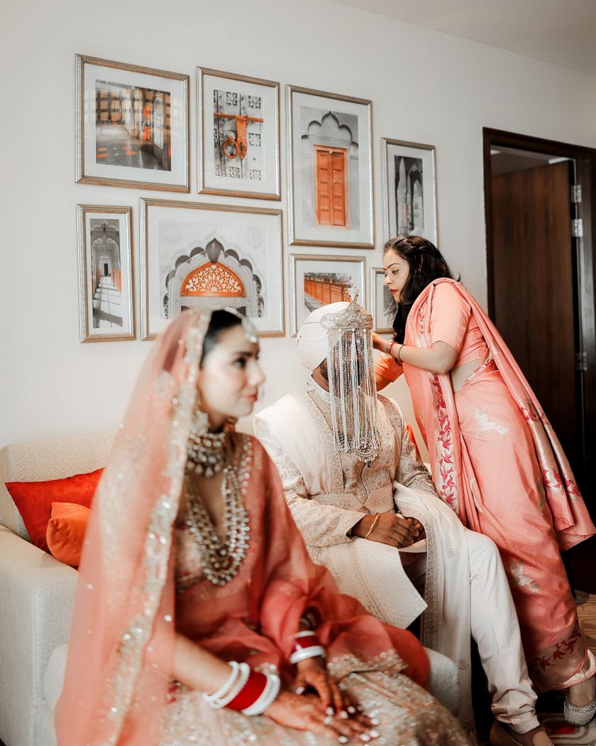 Cinematic wedding photography of a groom having his sehra adjusted while the bride sits softly blurred in the foreground, surrounded by family. | Studio Memory Lane